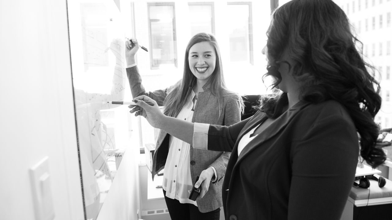 Two businesswomen discussing ideas and writing on a whiteboard in a bright office setting.