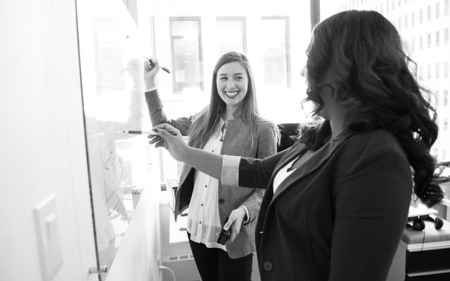 Two businesswomen discussing ideas and writing on a whiteboard in a bright office setting.