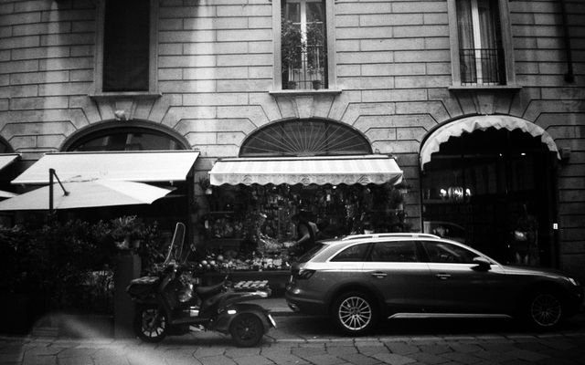 A monochrome urban street scene featuring a parked car, motor scooter, and a florist's storefront.