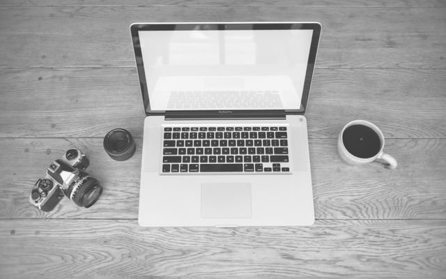 Monochrome image of a laptop, camera, lens, and coffee cup on a wooden desk