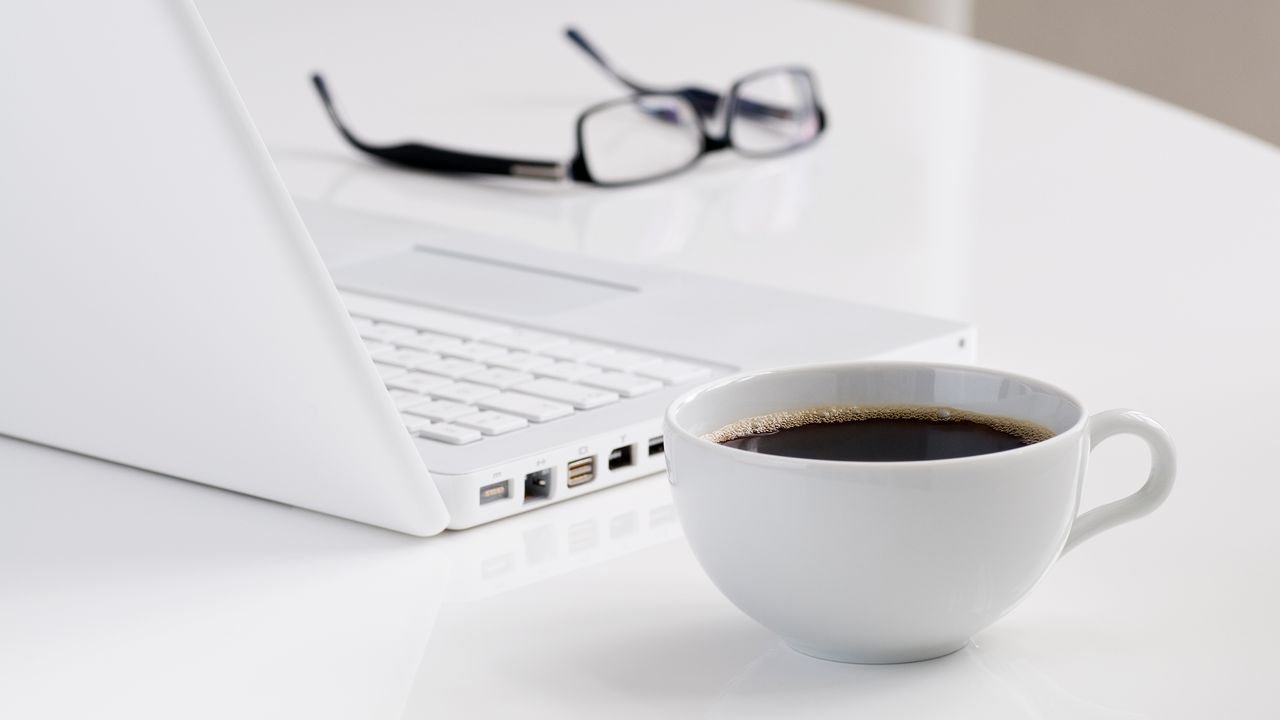 A minimalist workspace featuring a laptop, a cup of black coffee, and eyeglasses on a white desk.