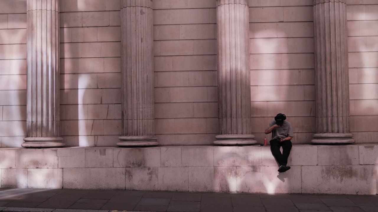 A lone figure sits against a historic columned wall in London, immersed in solitude.