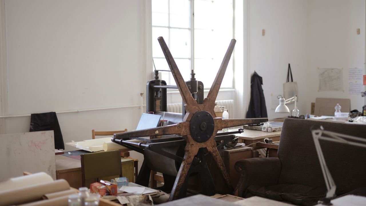 Workroom with paper rolls on table and white wall with notes and paper rolling machine with wooden mill shape wheel in afternoon