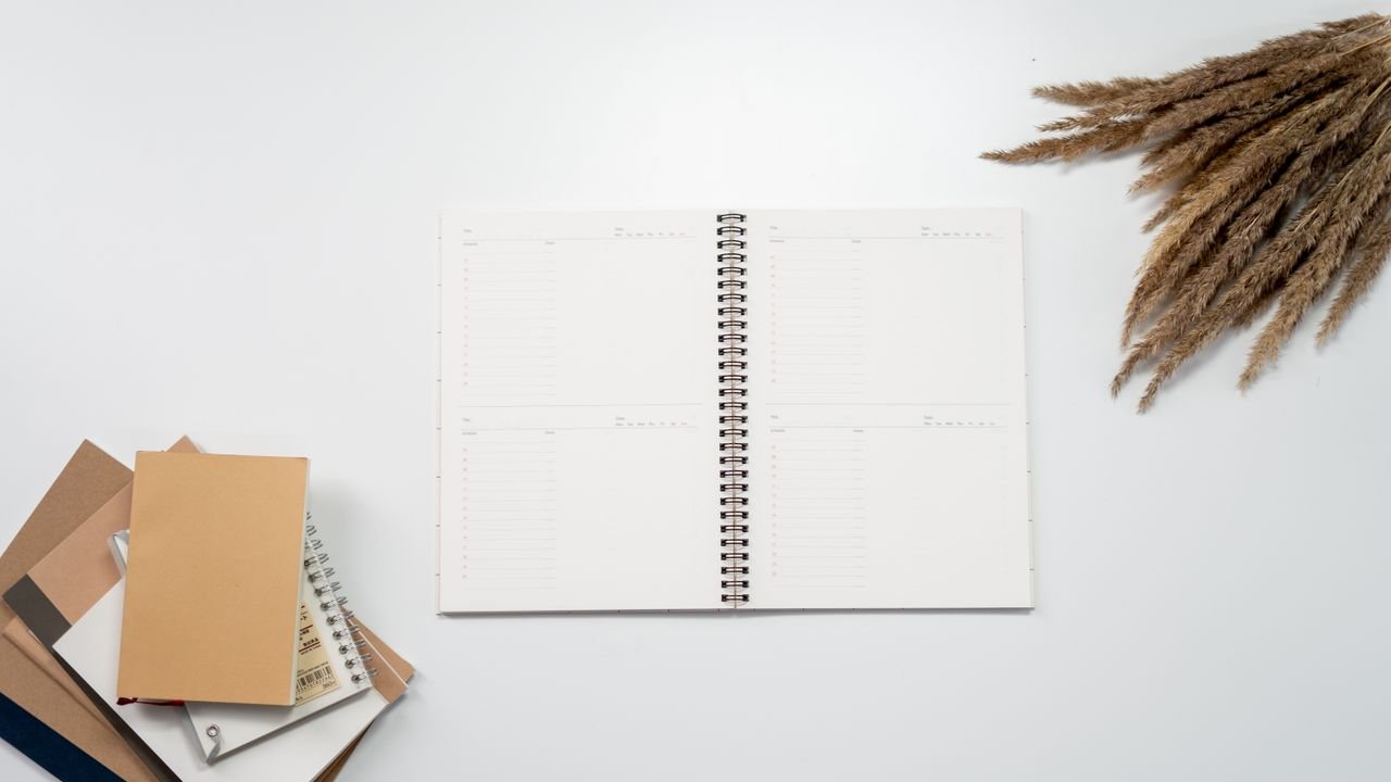 Open notebook with planner pages, surrounded by notebooks and pampas grass on a white background.
