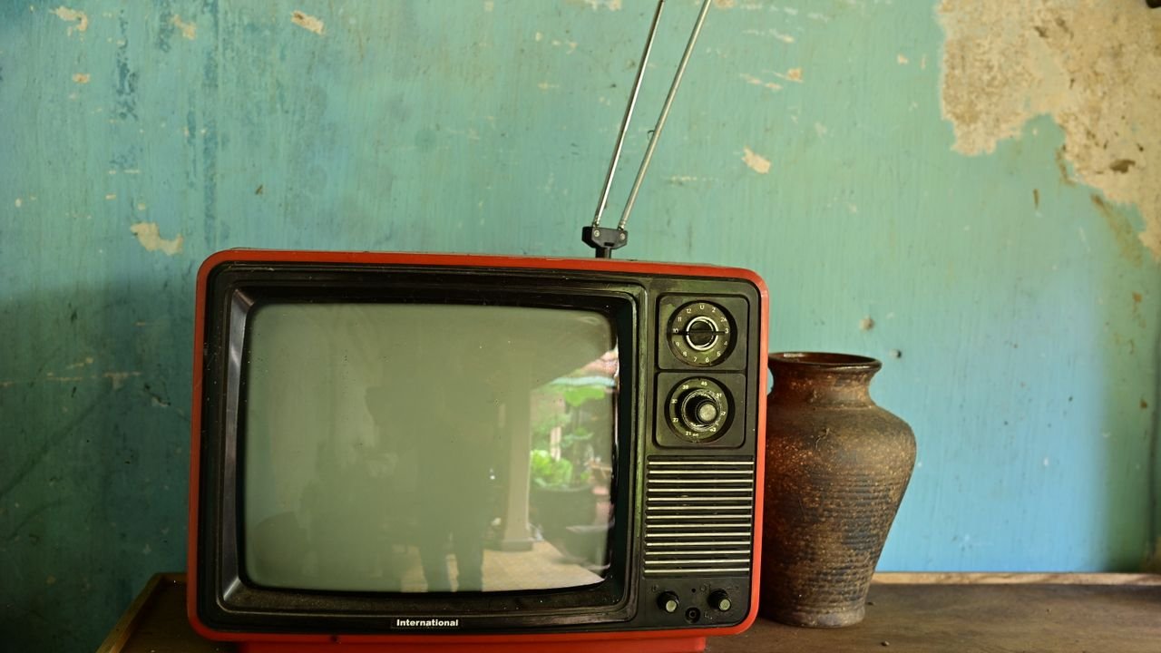 A vintage television with antenna beside a ceramic vase on a worn wooden surface against a rustic wall.