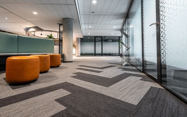 Spacious and modern office corridor featuring glass doors, geometric carpet patterns, and leather seating.