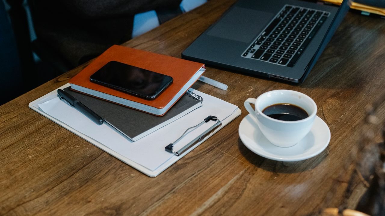 From above of opened laptop with cup of coffee placed on wooden table near notebooks and smartphone in office