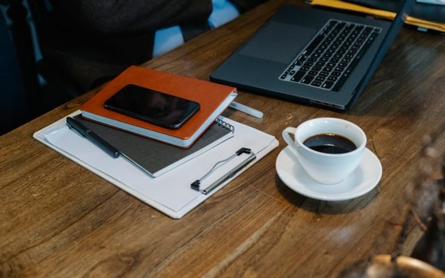 From above of opened laptop with cup of coffee placed on wooden table near notebooks and smartphone in office