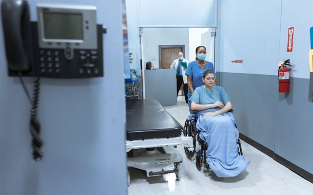 A nurse helps a patient in a wheelchair down a hospital corridor, reflecting care and medical professionalism.