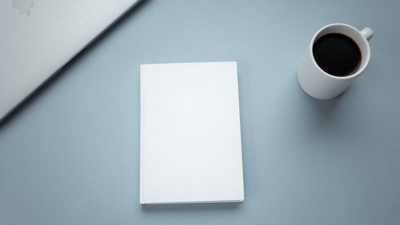 Top view of closed laptop placed near mug of hot americano and white hardcover book on table
