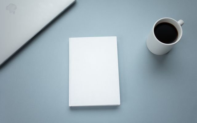 Top view of closed laptop placed near mug of hot americano and white hardcover book on table