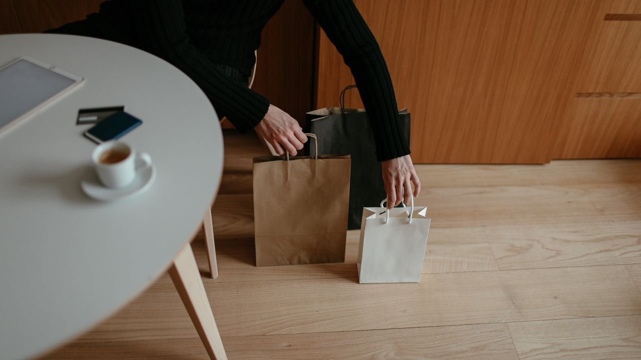 From above of crop unrecognizable male shopaholic in casual clothes sitting at table with tablet and coffee and putting paper bags on floor after shopping