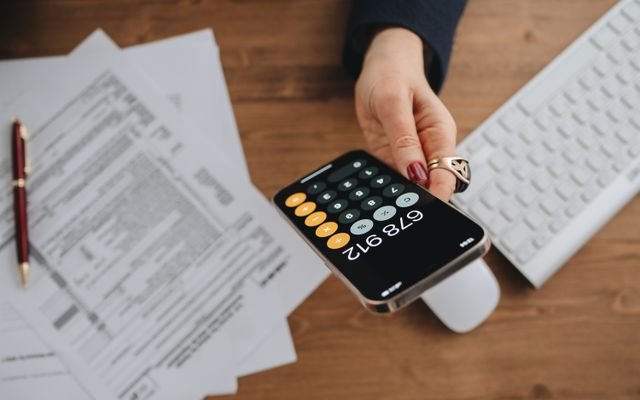 Hand holding smartphone calculator over tax documents on desk.