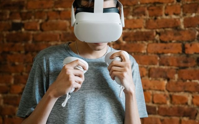 Young unrecognizable male in gray casual t shirt experiencing virtual reality with headset and controllers on background of brick wall