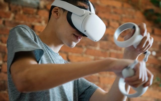 Crop young male in casual clothes using goggles of virtual reality and controllers while standing near brick wall