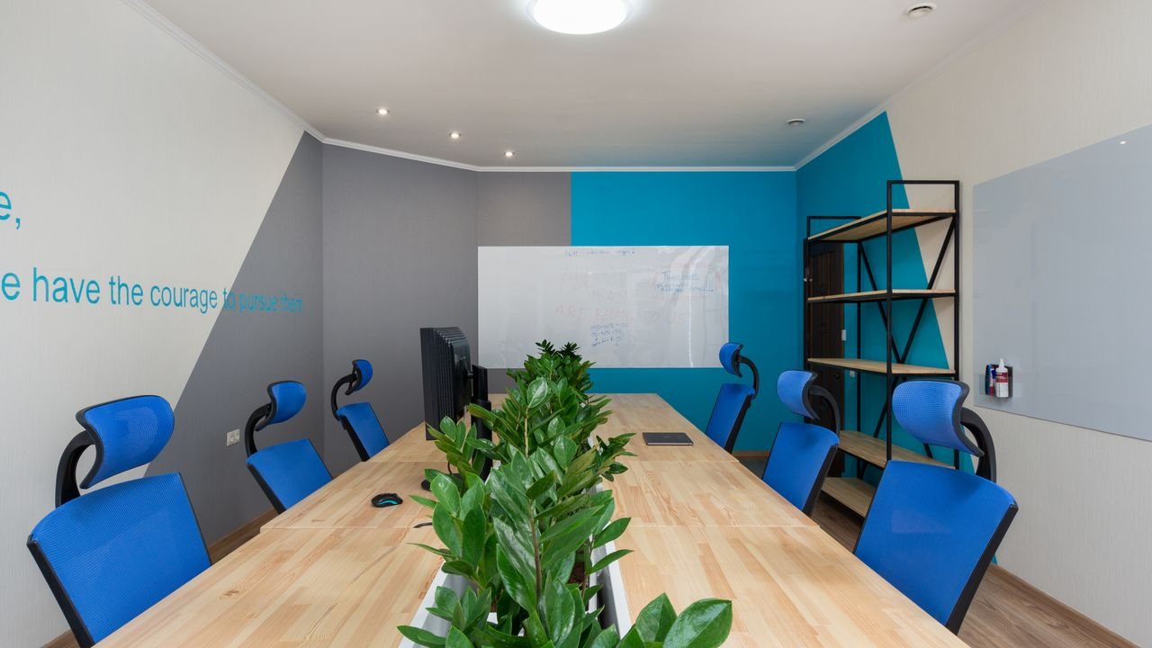 Armchairs at table with green plants placed in light conference room with inscription on wall and shelves in office building
