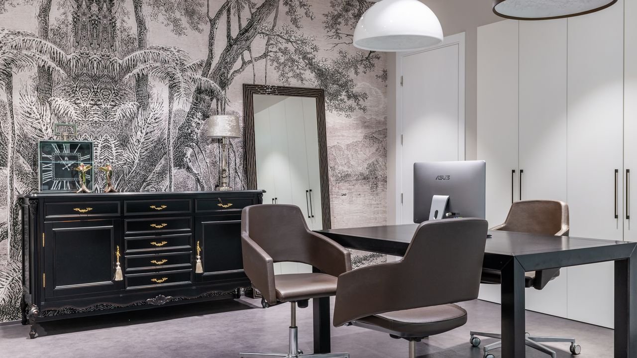 Modern computer on black table near armchairs placed in living room near cabinet with clock in spacious apartment with mirror and closet