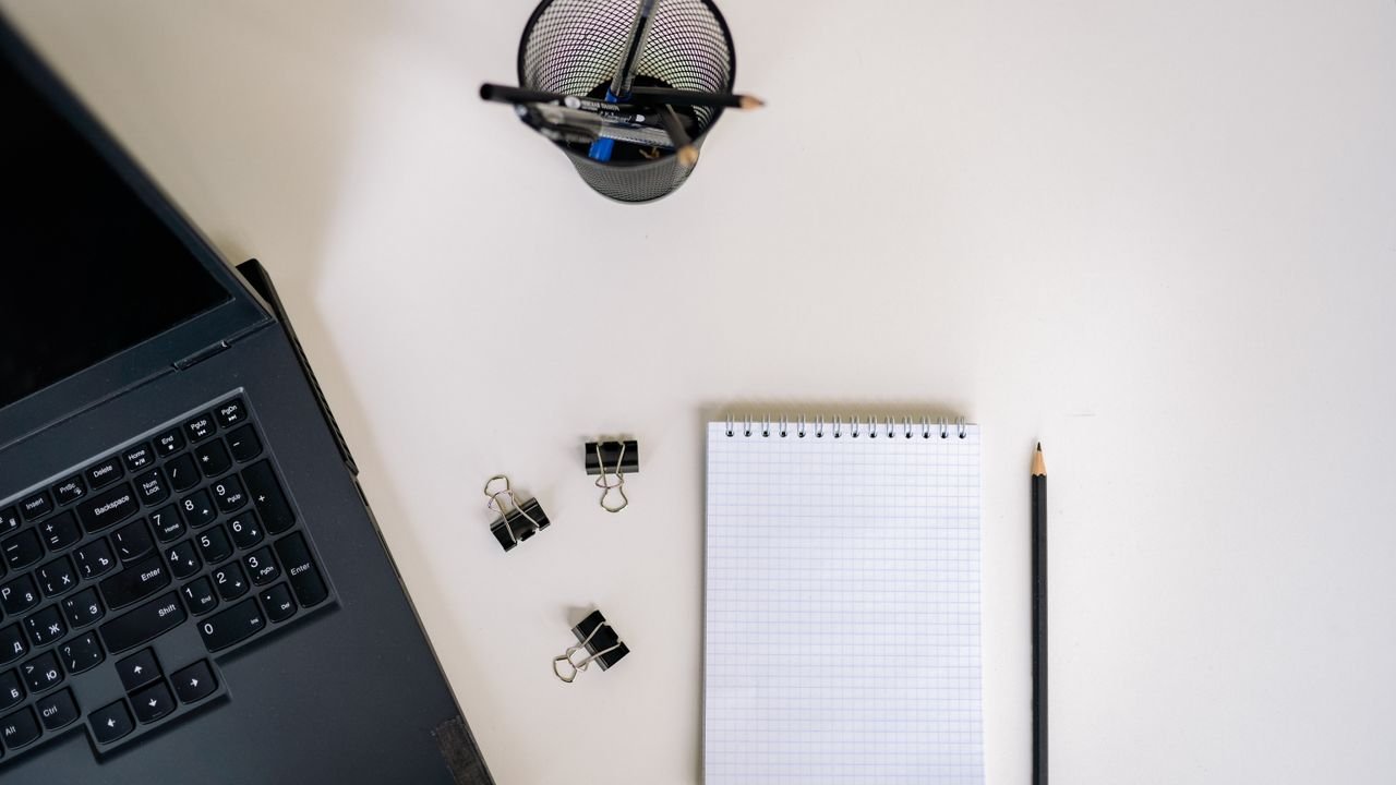 Top view of an office setup with a laptop, notepad, pencils, and clips on a white desk.