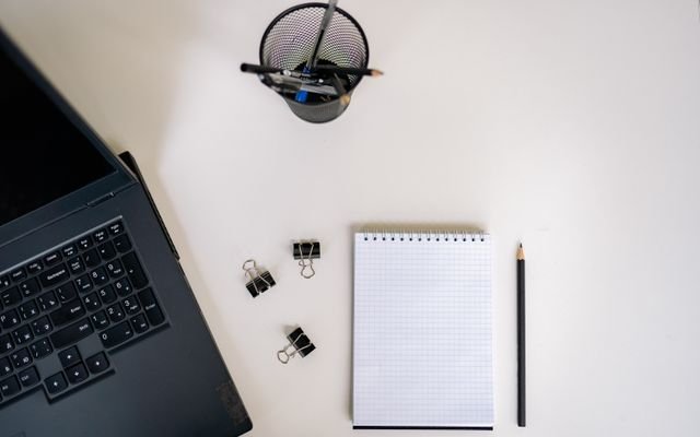 Top view of an office setup with a laptop, notepad, pencils, and clips on a white desk.